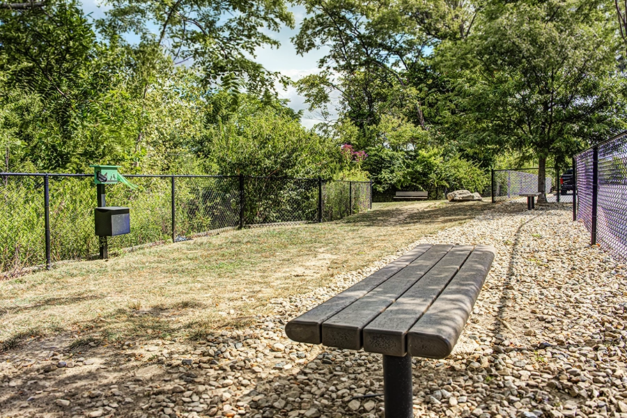 shaded fenced in area with a wooden bench during a bright sunny day. Pet waste disposable station against the fence.